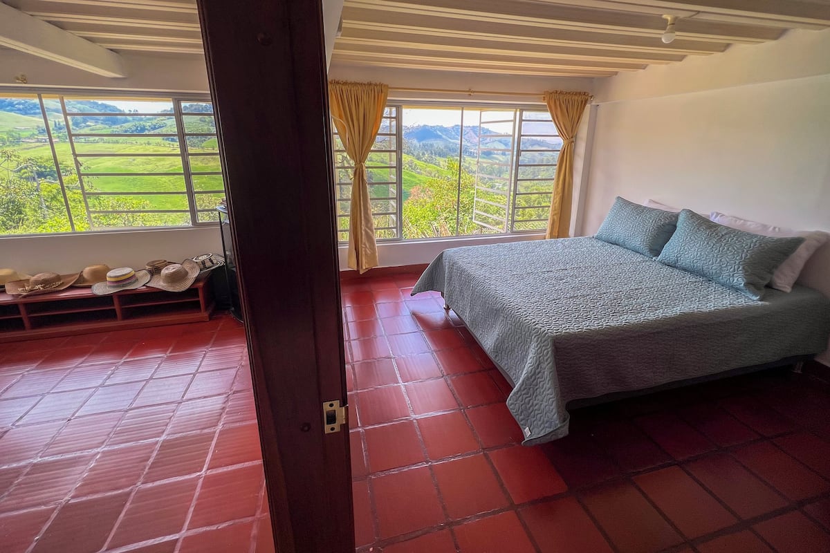 Une chambre à coucher avec un lit, un banc sur lequel sont posés des chapeaux, une fenêtre avec des rideaux et une vue sur la verdure.