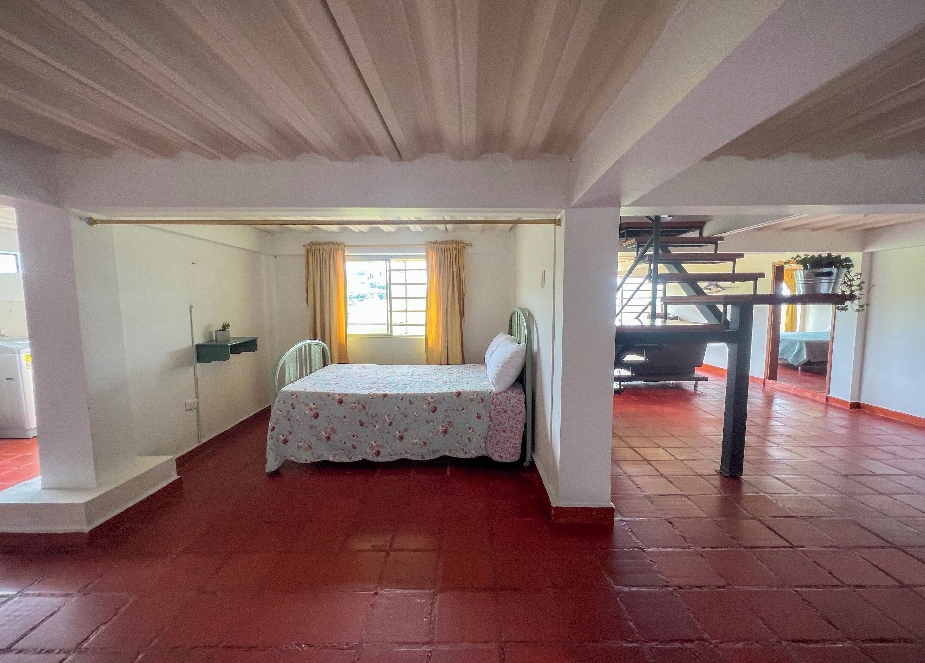 Une chambre à coucher avec un lit, une fenêtre avec des rideaux, une chaise et un escalier menant à une autre pièce.
