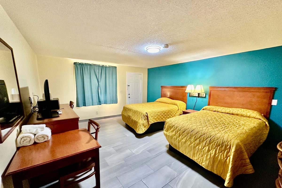 Quarto de hotel com duas camas, uma mesa, uma cadeira, uma televisão e uma janela com cortinas.