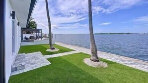A modern house with a patio overlooking a body of water, featuring a well-maintained lawn and two palm trees.