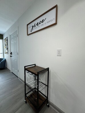 A room with a wooden floor, a white door, a wall-mounted sign with the word "grace" and a quote, and a metal cart with wine glasses. - DT Dallas 1BD BOHO Abode 5 Min from Love Field (Dallas)