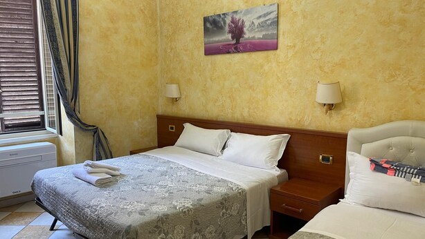 A hotel room with a bed, a nightstand, a bedside table, a window with blinds, a wall-mounted lamp, and a painting. - HOLIDAY ROMA (Rome)