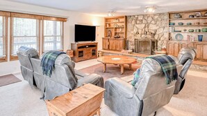 A living room with leather sofas, a wooden coffee table, a flat-screen TV, a stone fireplace, and shelves with decorative items. - Fireplace | Pool Table | WiFi | Hot Tub | Deer Haven (Angel Fire)