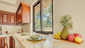 A kitchen with wooden cabinets, a marble countertop, and a window with a view of greenery. - NOUR Villas (Belmopan)