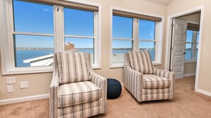 Una habitación con dos sillones a rayas, un otomano redondo oscuro y grandes ventanas con vistas a un cuerpo de agua.