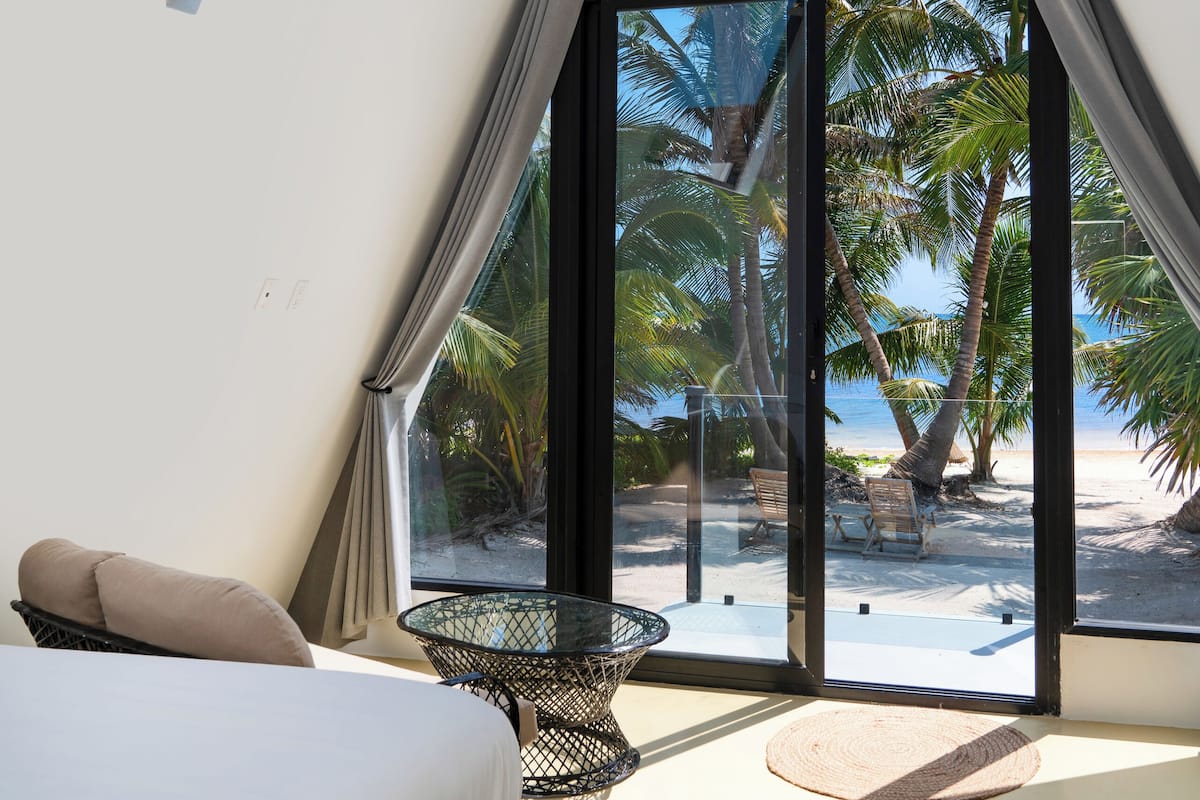 A modern room with a bed, a ceiling fan, a glass door leading to a beach view, and a small table.