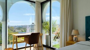 Une chambre d’hôtel avec une grande fenêtre, un bureau, une chaise, un lit et une table de chevet avec une lampe.