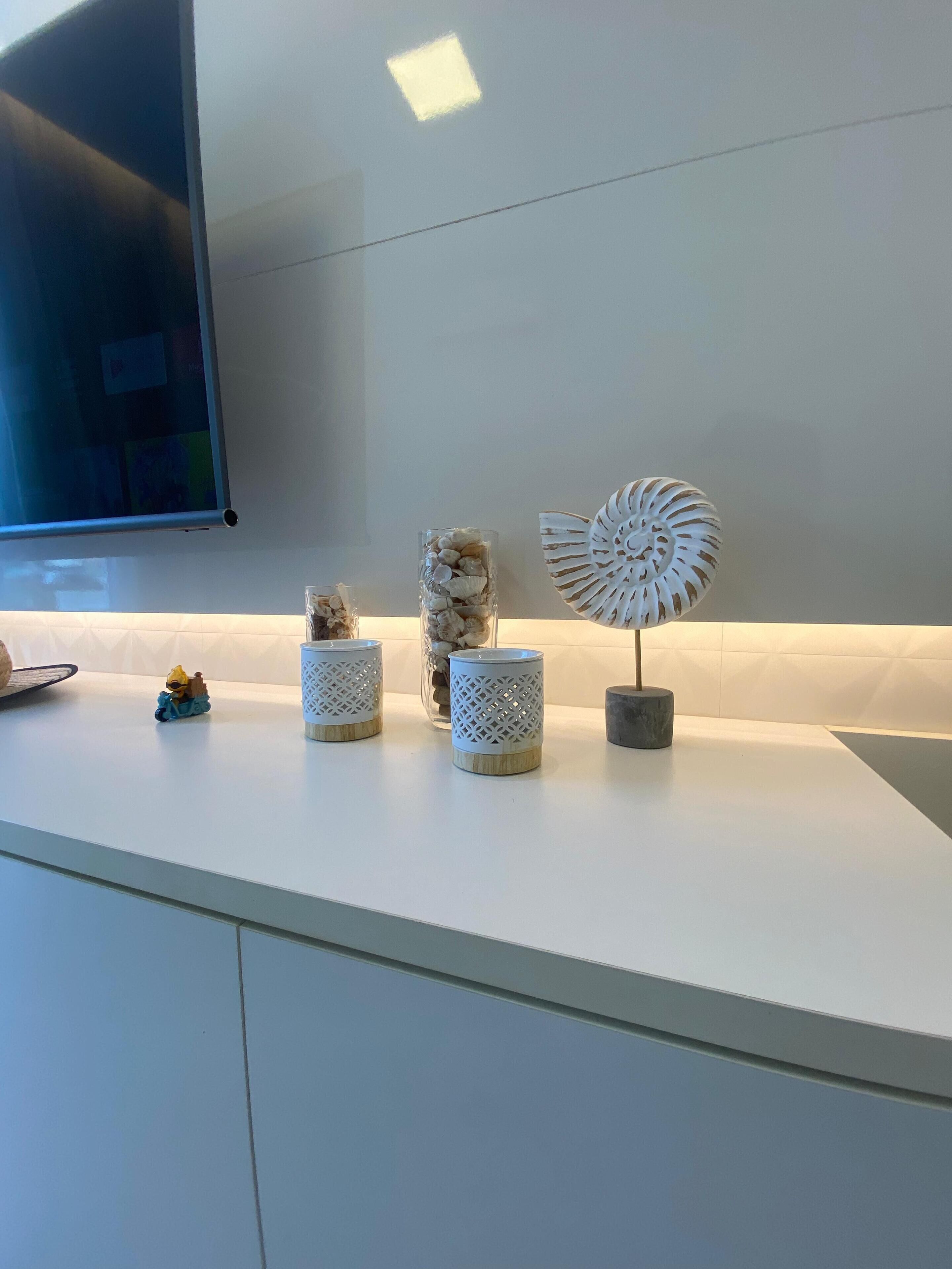 A modern kitchen with white cabinets, a flat-screen TV mounted on the wall, and decorative items including a small figurine, two glass containers with shells, and a white decorative piece.