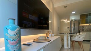 A modern kitchen with a large flat-screen TV mounted on the wall, a white countertop, and a dining area with white chairs. - Flat 110 Muro Alto CC by LaPlaya (Ipojuca)