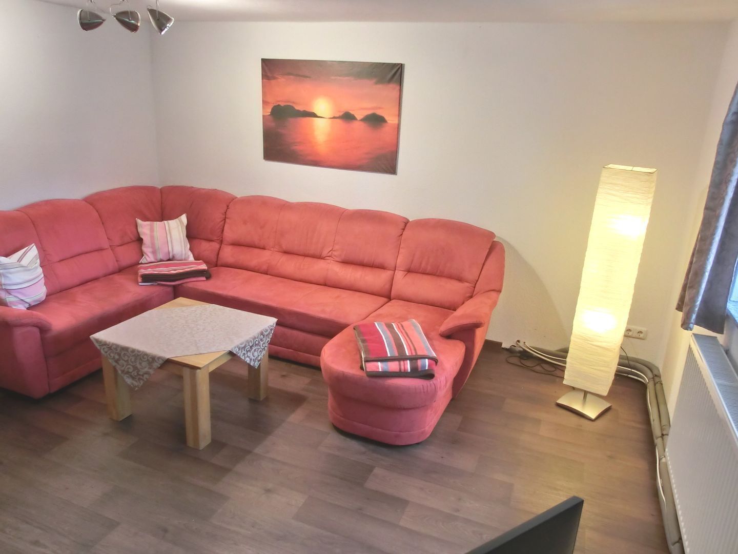 A living room with a pink sofa, a small table, a floor lamp, and a painting of a sunset.