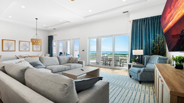 A spacious living room with a large grey sofa, a striped armchair, and a wooden coffee table. There is a balcony with a view of the sea.