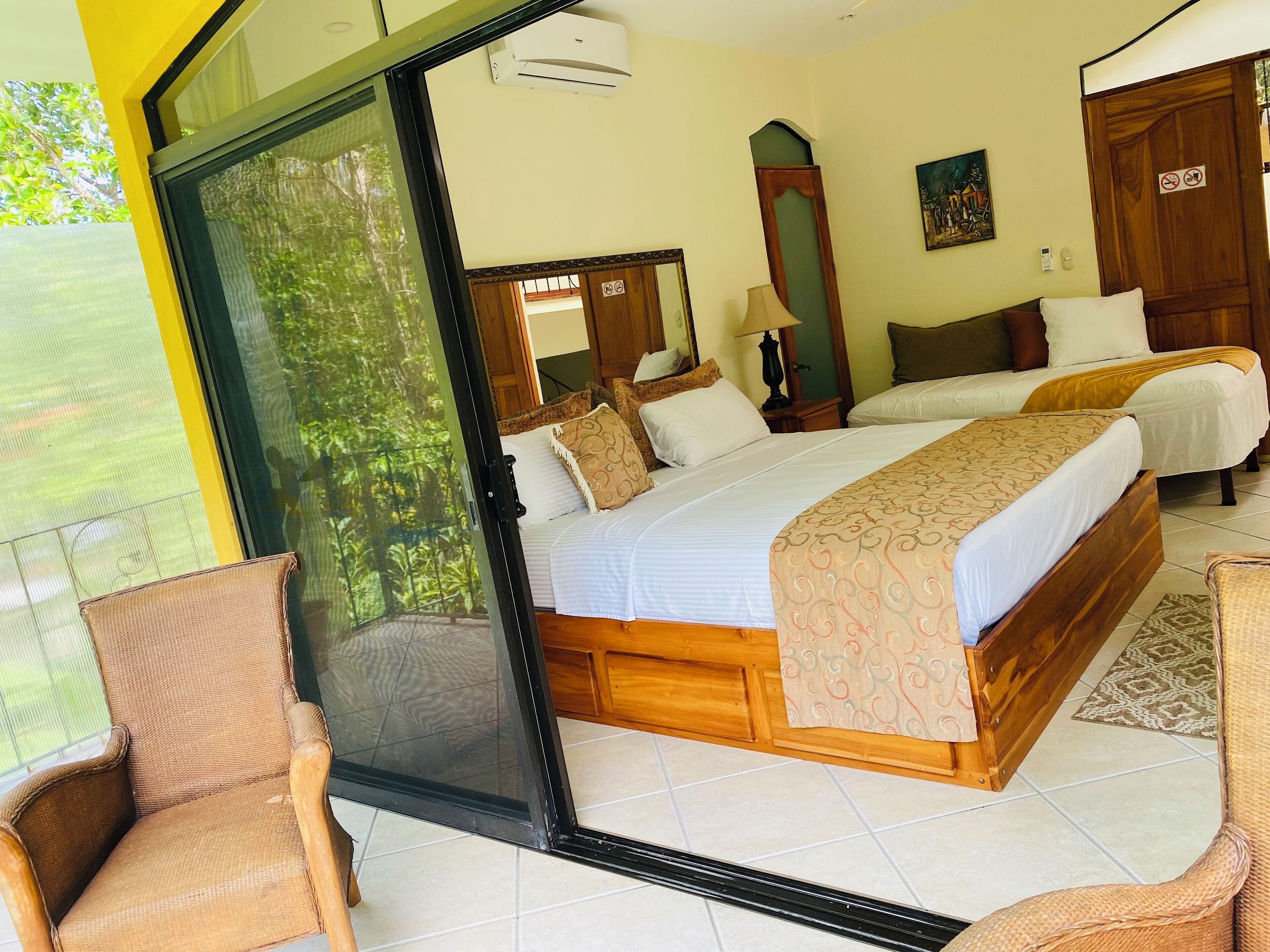 A bedroom with a large bed, two bedside tables, a mirror, and a window with a view of greenery.