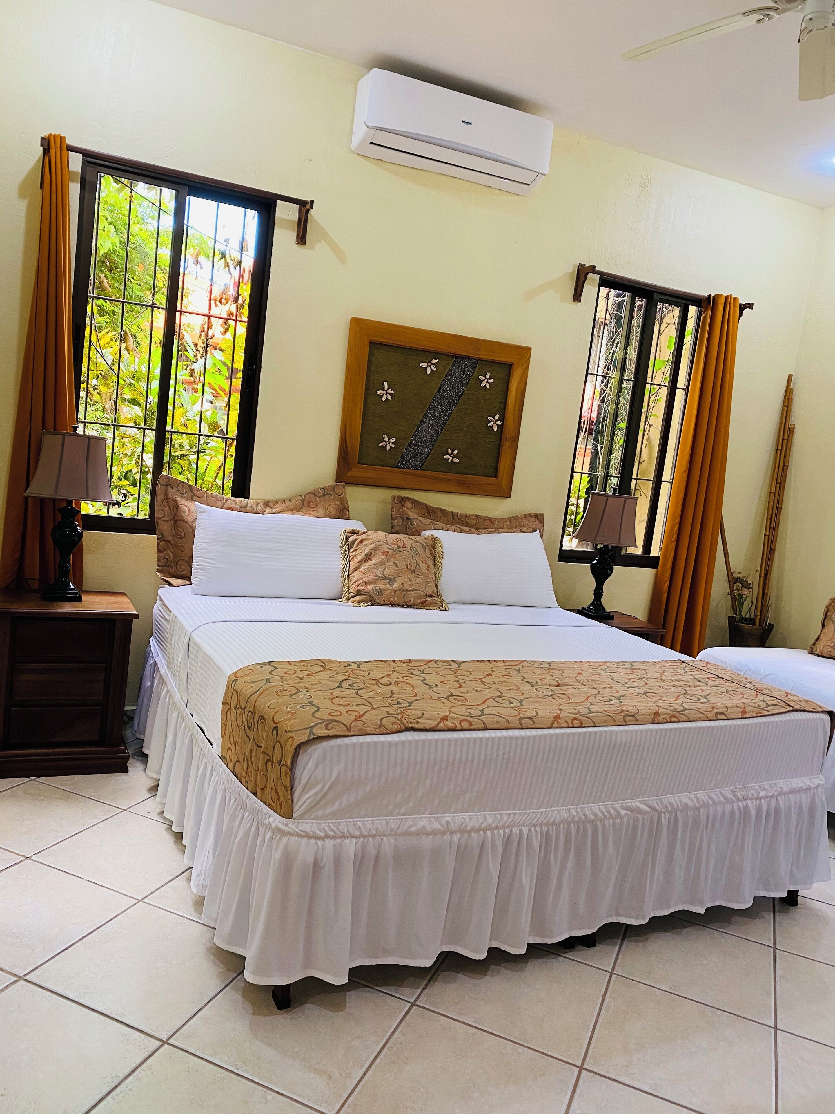 A hotel room with a large bed, two bedside tables, a window with curtains, an air conditioning unit, and a framed picture on the wall.