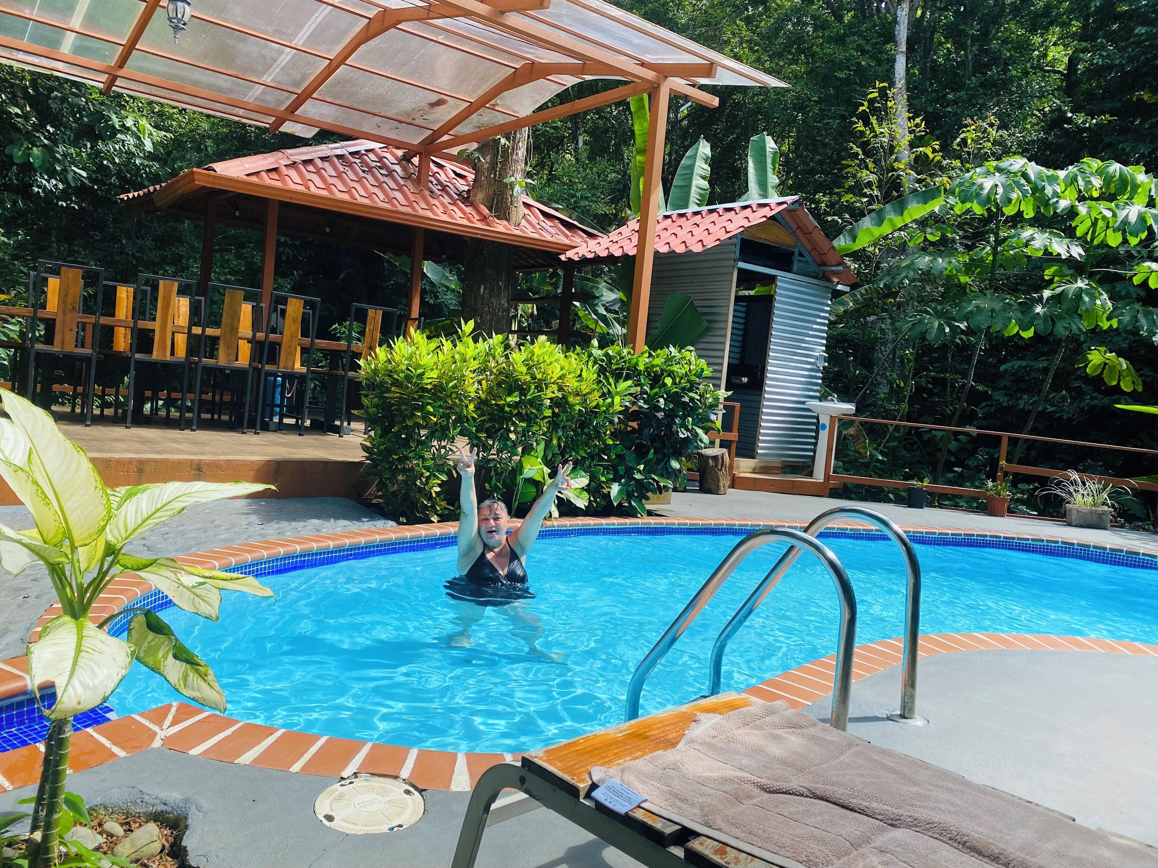 Une piscine avec un bord carrelé, une terrasse en bois et un espace de détente couvert équipé de chaises.