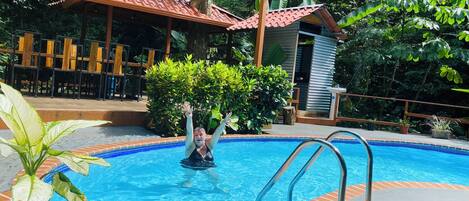 Une piscine avec un bord carrelé, une terrasse en bois et un espace de détente couvert équipé de chaises.