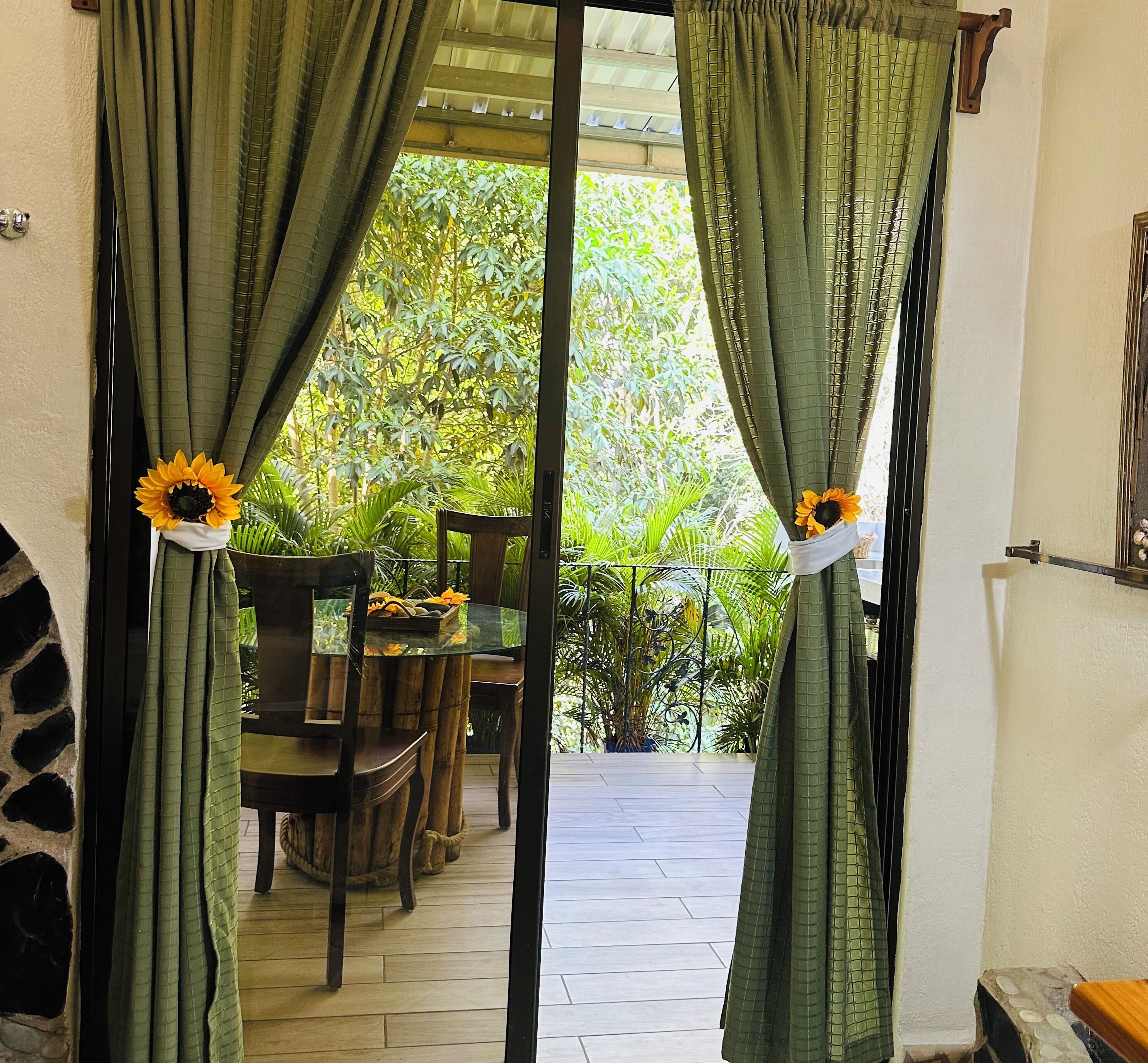 A room with a glass door leading to an outdoor area with a dining table and chairs, surrounded by lush greenery.