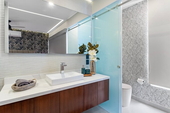 Un baño moderno con un lavamanos blanco, muebles de madera, cerramiento de ducha de vidrio y un jarrón decorativo con flores.