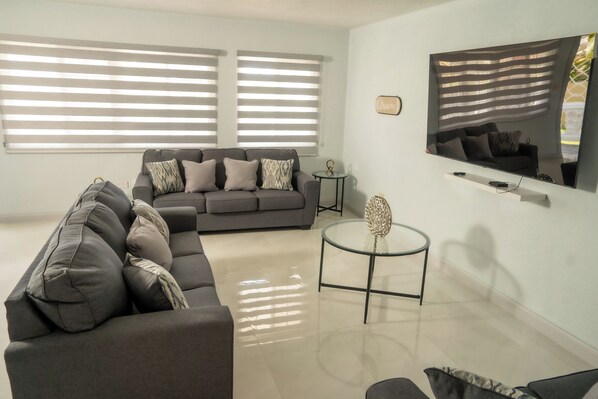A modern living room with a grey sofa, a glass coffee table, and a flat-screen TV mounted on the wall. - North Ritz Villa - 1 Bedroom Apartment (Ocho Rios, Draxhall, St Ann, Jamaica)