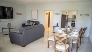 A modern living room with a grey sofa, a dining table set with plates and glasses, and a kitchen in the background. - North Ritz Villa - 1 Bedroom Apartment (Ocho Rios, Draxhall, St Ann, Jamaica)