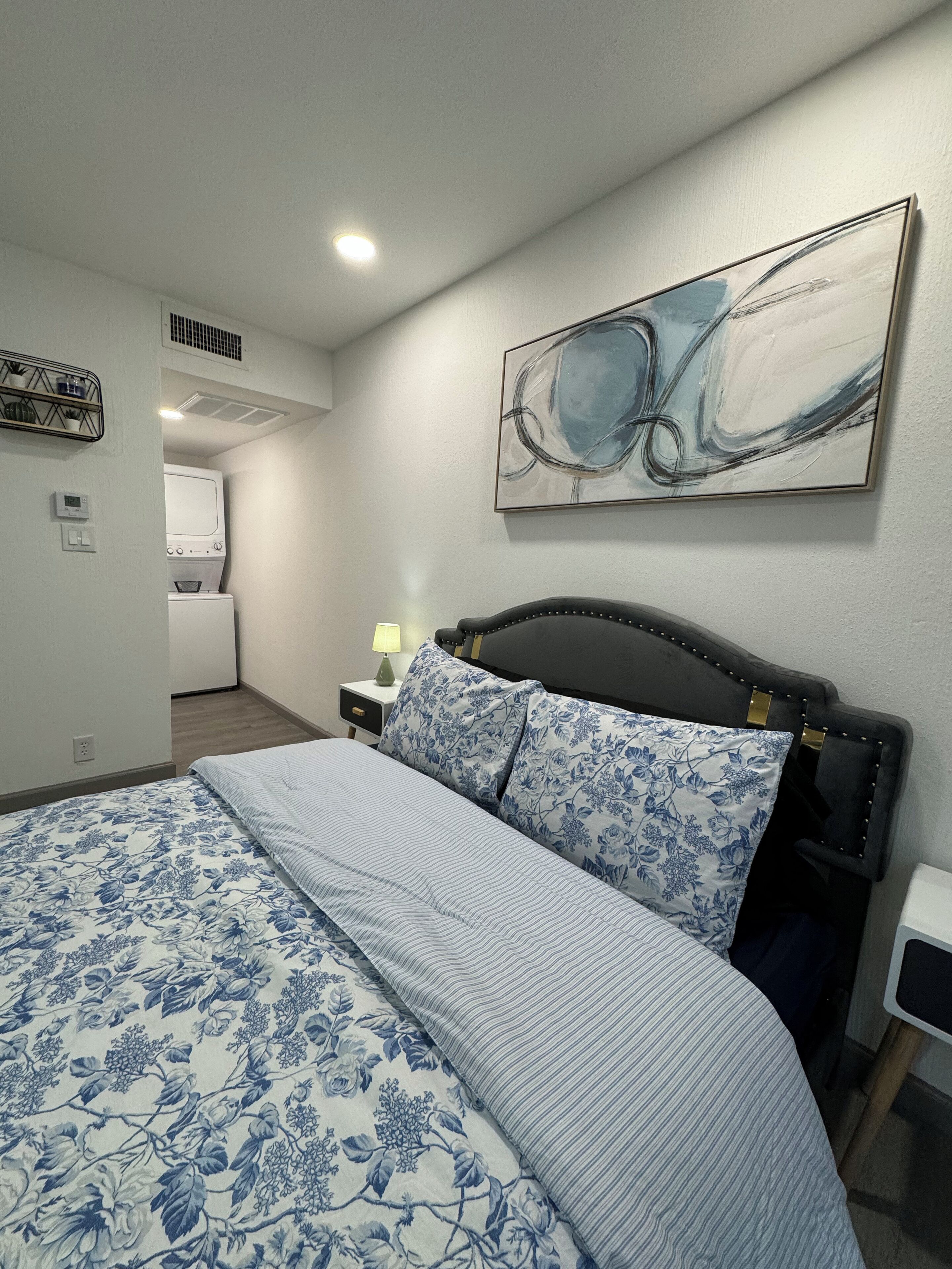 Ein Schlafzimmer mit einem Bett, einem Nachttisch, einer Lampe, einem Wandbild im abstrakten Stil und einem Wäschebereich im Hintergrund.