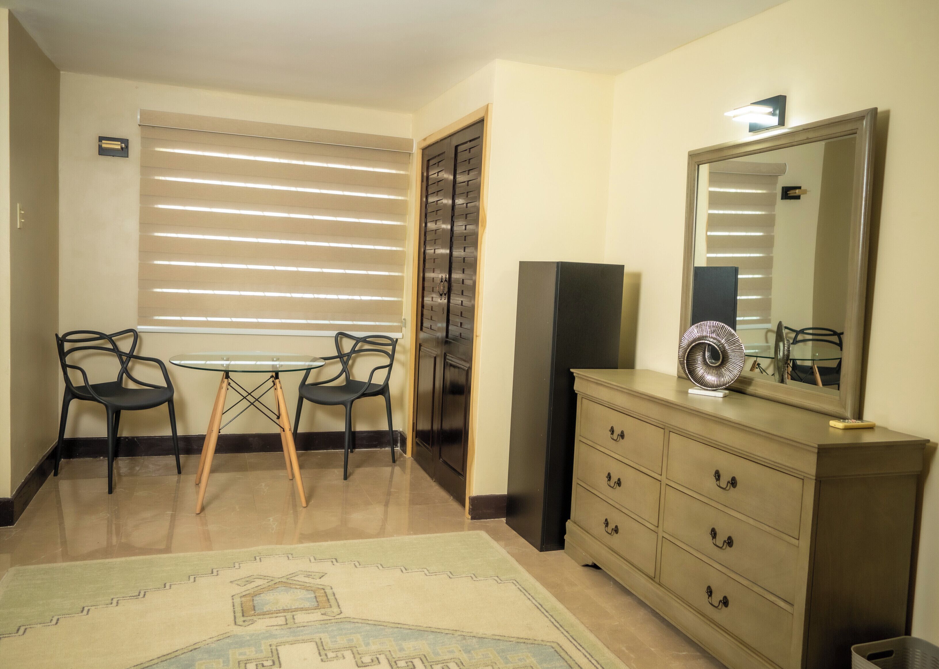 Una habitación con una mesa de comedor, dos sillas, un aparador, un espejo y una ventana con persianas.