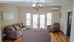 A living room with a large sofa, two armchairs, a coffee table, and a chandelier. There is a door with glass panels and an air conditioning unit above it.