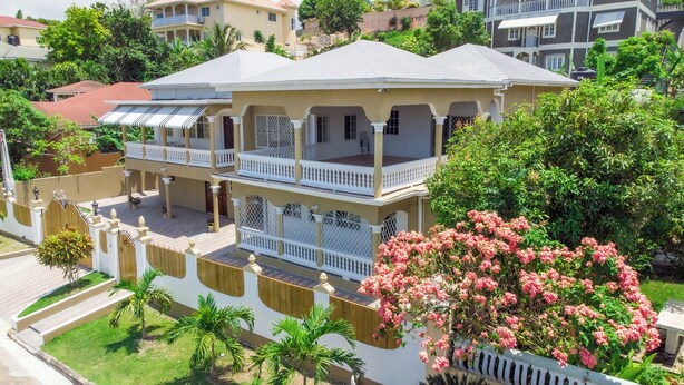 A multi-story house with a balcony, surrounded by greenery and other houses.