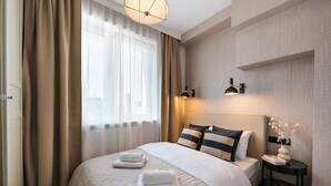 Ein Hotelzimmer mit einem großen Bett, einem Nachttisch mit Lampe und Blick auf die Stadt durch das Fenster.