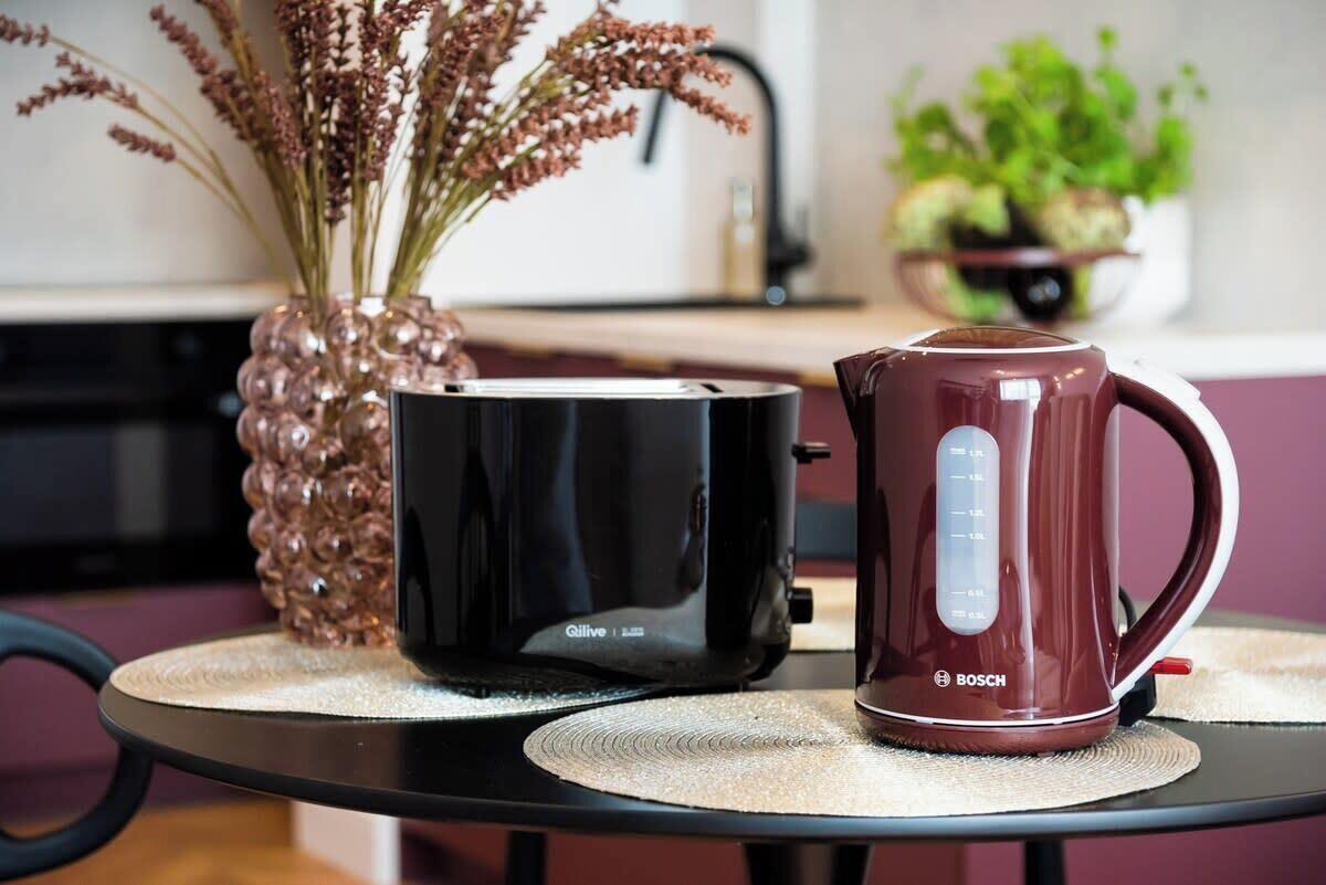 A kitchen with a black toaster, a maroon coffee maker, and a vase with dried plants on a table.