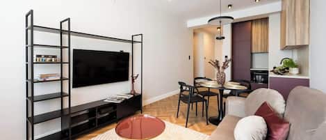 A modern living room with a flat-screen TV, a black entertainment unit, a round coffee table, a beige sofa with cushions, a dining area with chairs, and a pendant light.