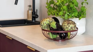 Eine Küchenarbeitsplatte mit Marmorplatte, einem schwarzen Wasserhahn, einem weißen Topf mit Grünpflanzen und einem Drahtkorb mit Artischocken und einer Aubergine.