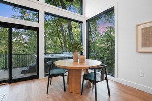 A modern dining area with a round wooden table, two chairs, and a vase. Large windows offer a view of trees and a deck outside.