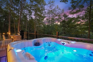 A hot tub on an outdoor deck with a wooden railing, surrounded by trees and a twilight sky.