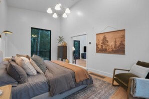 A modern bedroom with a large bed, a view of the forest through a window, and a painting on the wall.