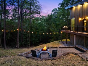 Eine moderne Terrassengruppe im Freien mit Feuerstelle, umgeben von Stühlen und Lichterketten, vor dem Hintergrund hoher Bäume und eines Holzgebäudes mit Balkon.