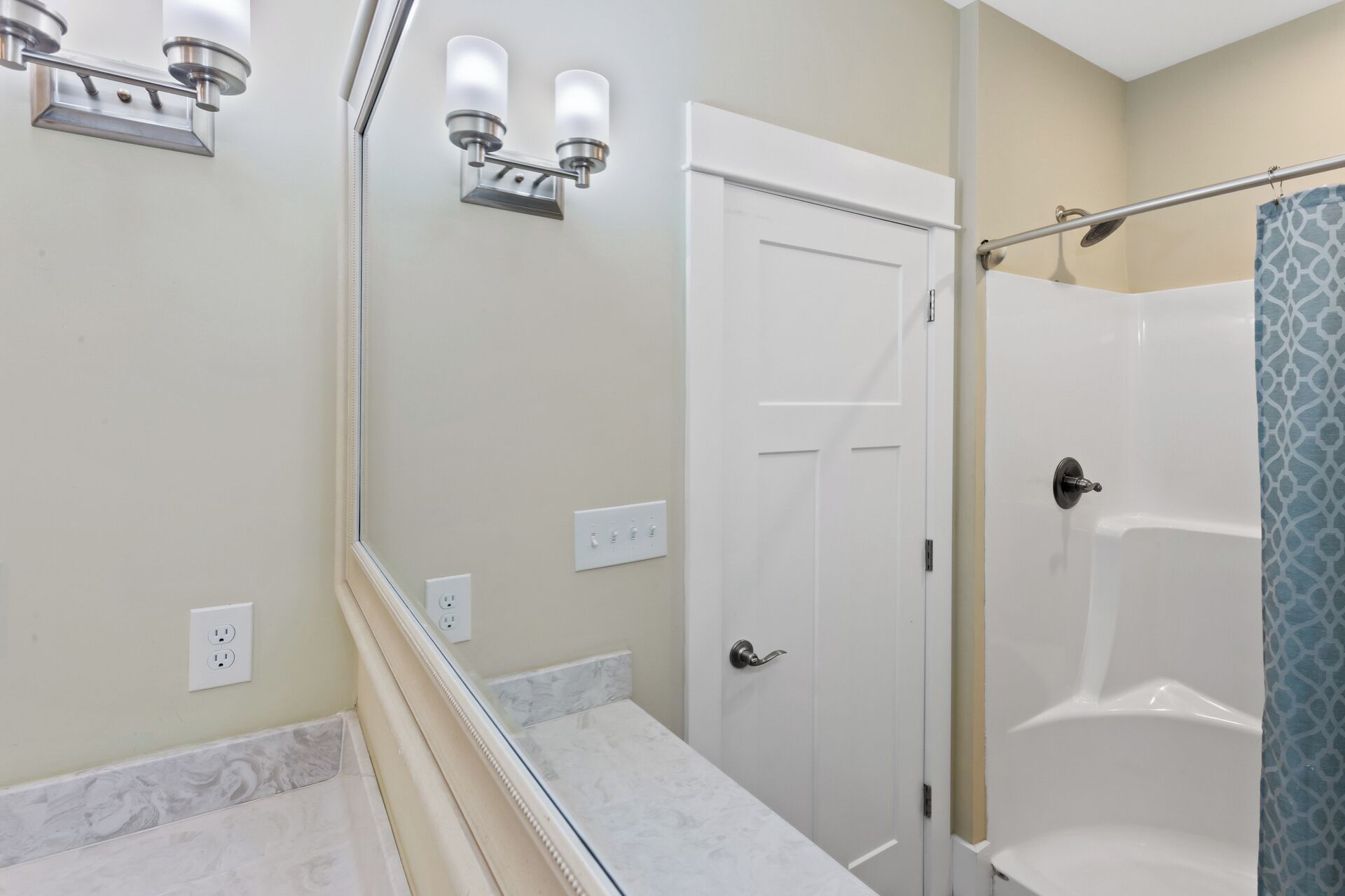 Une salle de bain avec une porte blanche, une douche avec une porte en verre et un miroir surmonté d’un luminaire.