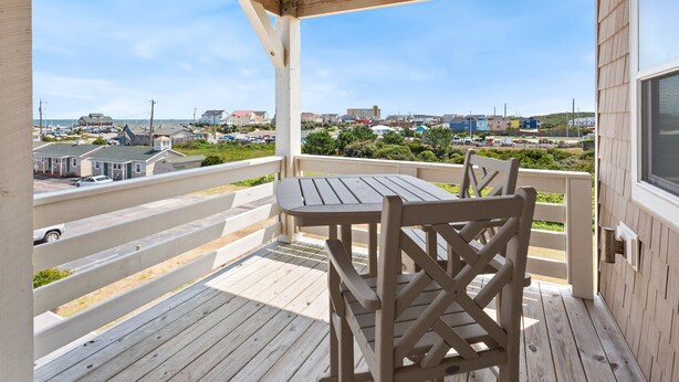 Una terraza de madera con una mesa y sillas, con vistas a una zona costera con edificios y un estacionamiento.
