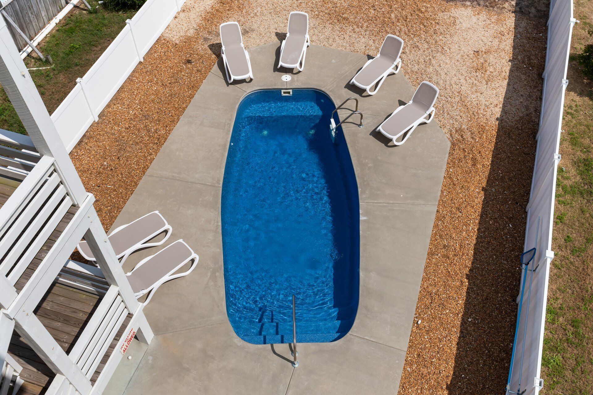 Une piscine rectangulaire avec un escalier en métal, entourée de chaises longues blanches et d’une terrasse en bois.