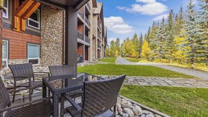 Un bâtiment moderne à l’extérieur en pierre et en bois, doté d’un espace extérieur aménagé avec des chaises en osier et offrant une vue sur une zone boisée.