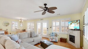 A living room with a sofa, a coffee table, a television, a fireplace, and a ceiling fan. - Shorely Bliss 3-Min Walk to Beach w/ Hot Tub (Kitty Hawk)