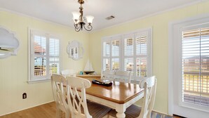 A dining area with a wooden table, white chairs, a chandelier, and large windows with shutters. - Shorely Bliss 3-Min Walk to Beach w/ Hot Tub (Kitty Hawk)