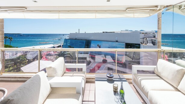 A balcony with white sofas and a table, overlooking a marina with boats and a building with a blue facade.