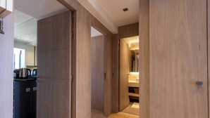 A modern hotel corridor with wooden flooring and doors leading to a bathroom and a kitchenette area.