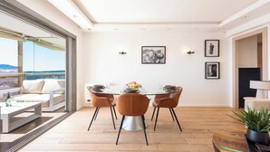 A modern dining area with a glass table, four brown chairs, and a view of the sea through large windows.