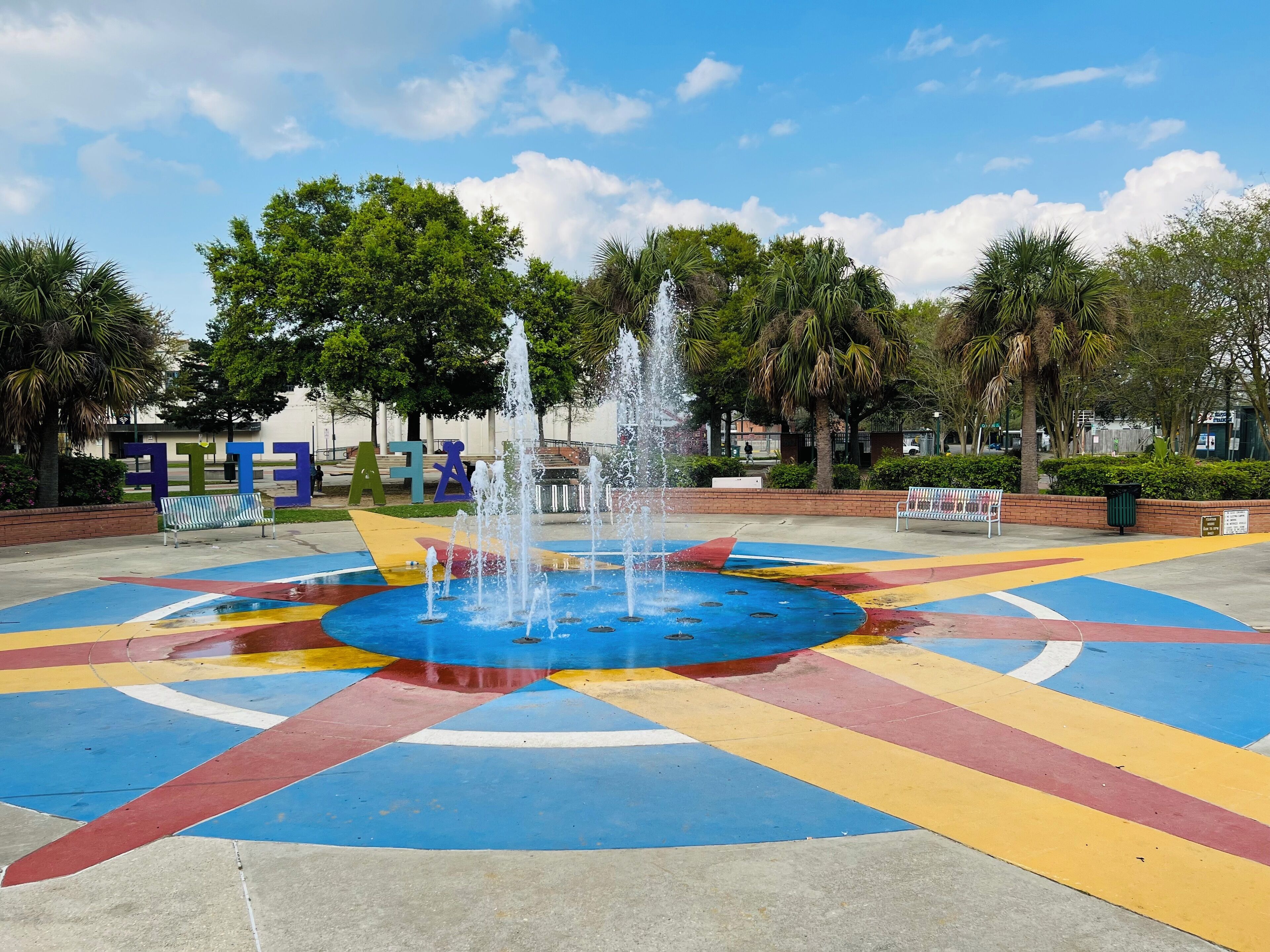A fountain with water jets in the center of a colorful tiled area, surrounded by benches and trees.