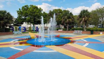 A fountain with water jets in the center of a colorful tiled area, surrounded by benches and trees.