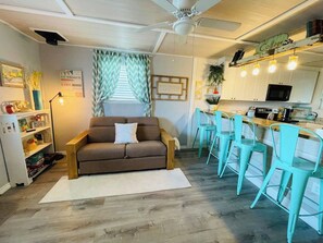 Un salon chaleureux avec un canapé marron, des fauteuils turquoise et un coin cuisine doté de meubles blancs et d’un four à micro-ondes.