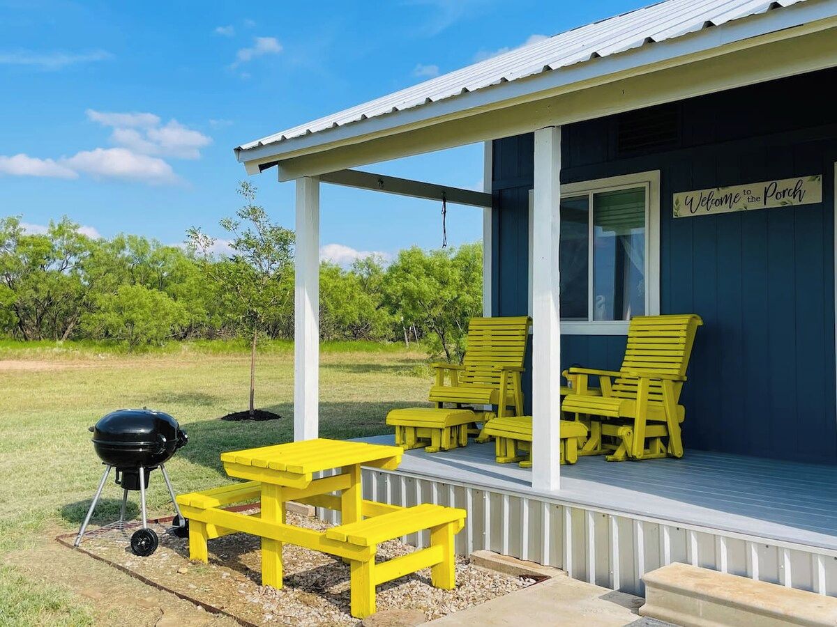Un porche con muebles de exterior amarillos, una parrilla de barbacoa y un letrero que dice 'Welcome to the Porch'.