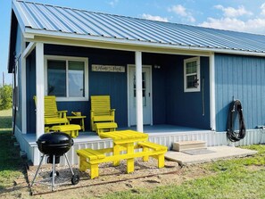 Un petit bâtiment bleu avec un porche, un barbecue et des meubles d’extérieur jaunes.