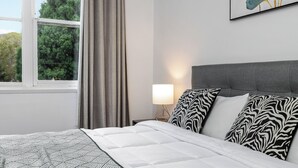 A neatly made bed with patterned bedding, a bedside lamp, and a window with curtains. - Cosy & Convenient Place in Inner West Area (no parking) (Strathfield)
